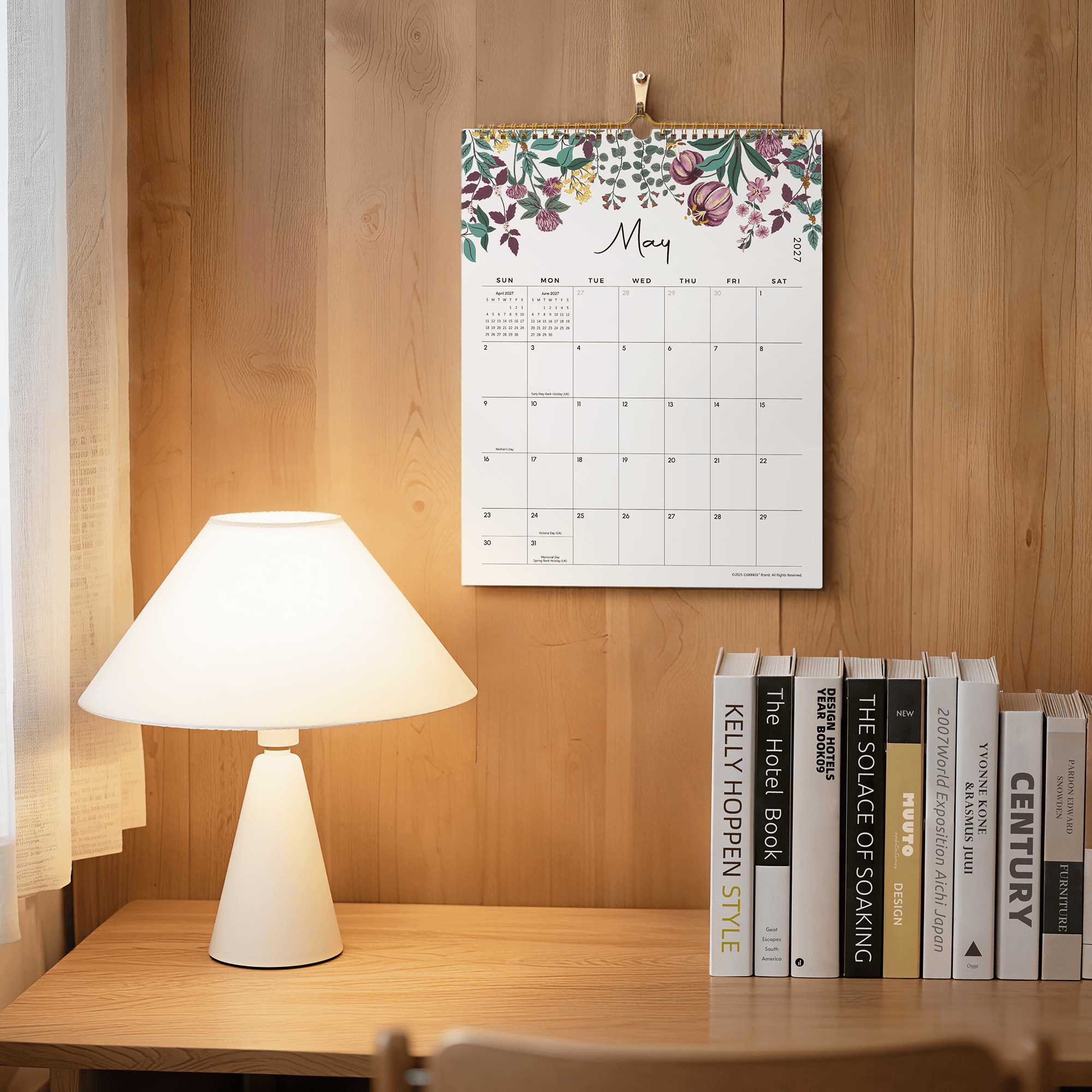 Floral calendar on a wooden wall above a table with books and a lamp.