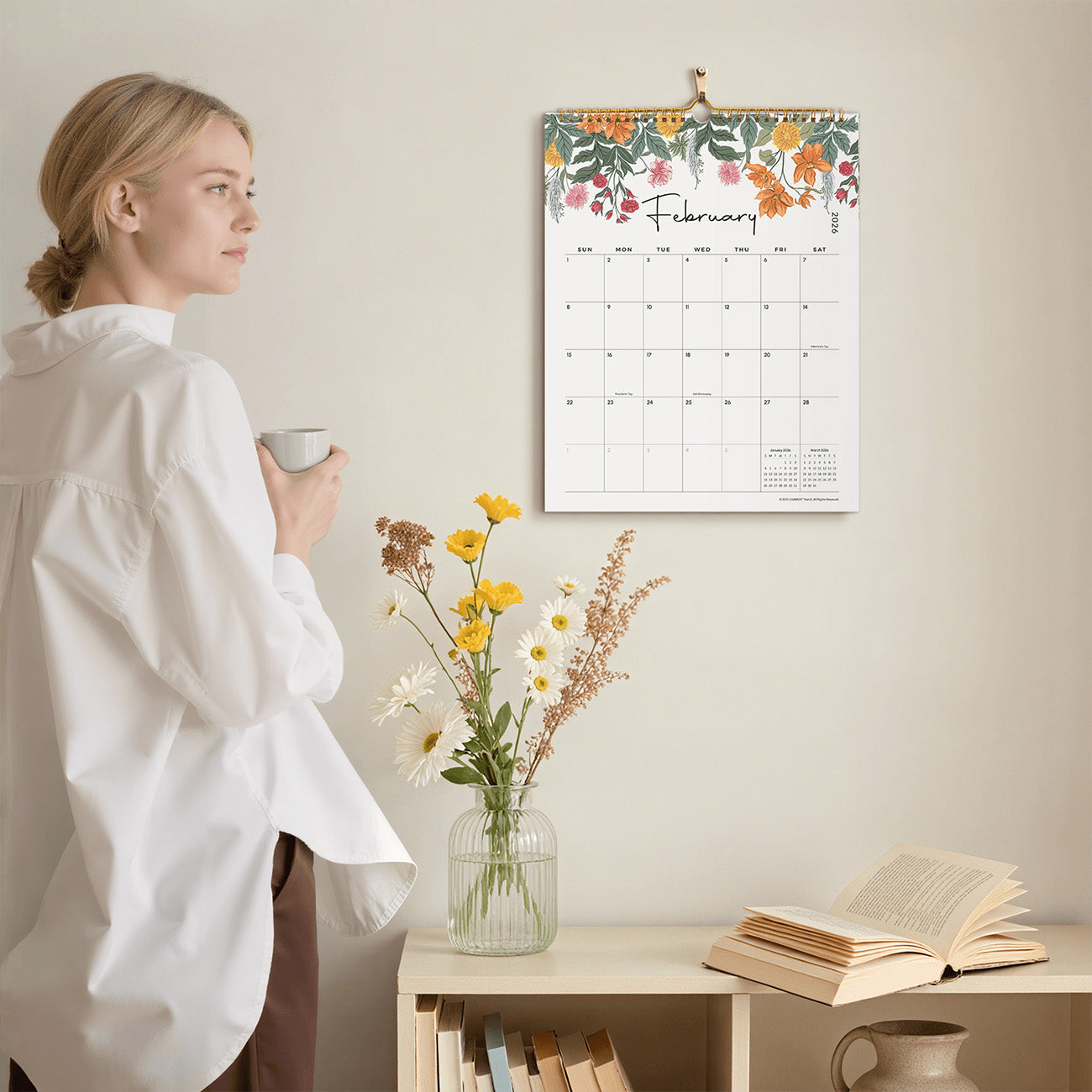 Woman holding a mug with a floral calendar on the wall, books, and a vase of flowers on a table.