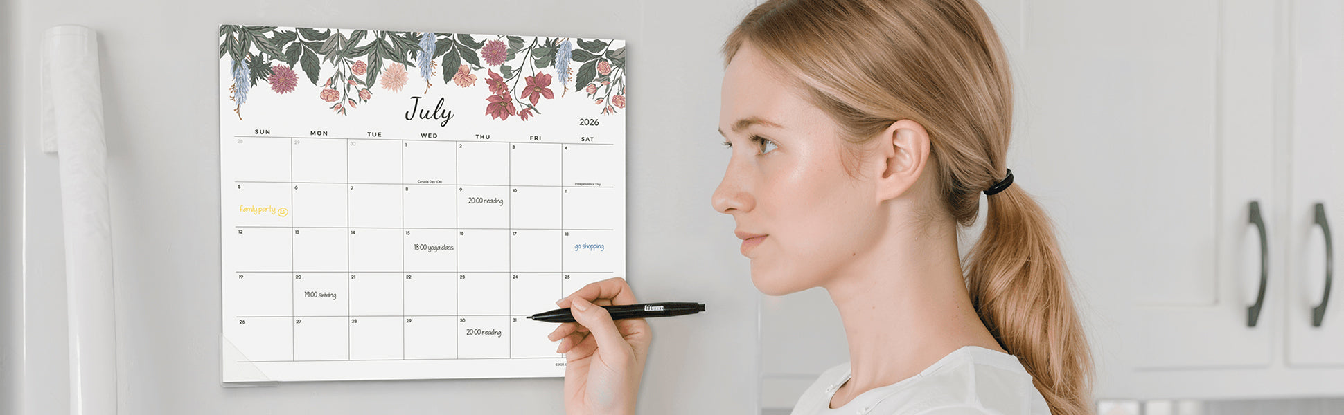 Woman holding a pen next to a floral July calendar on a wall.