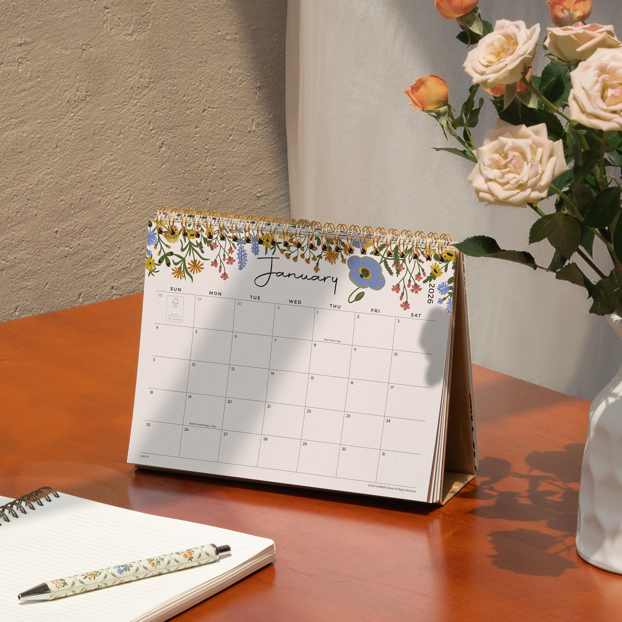 Floral desk calendar on a wooden surface with a pen and notebook.