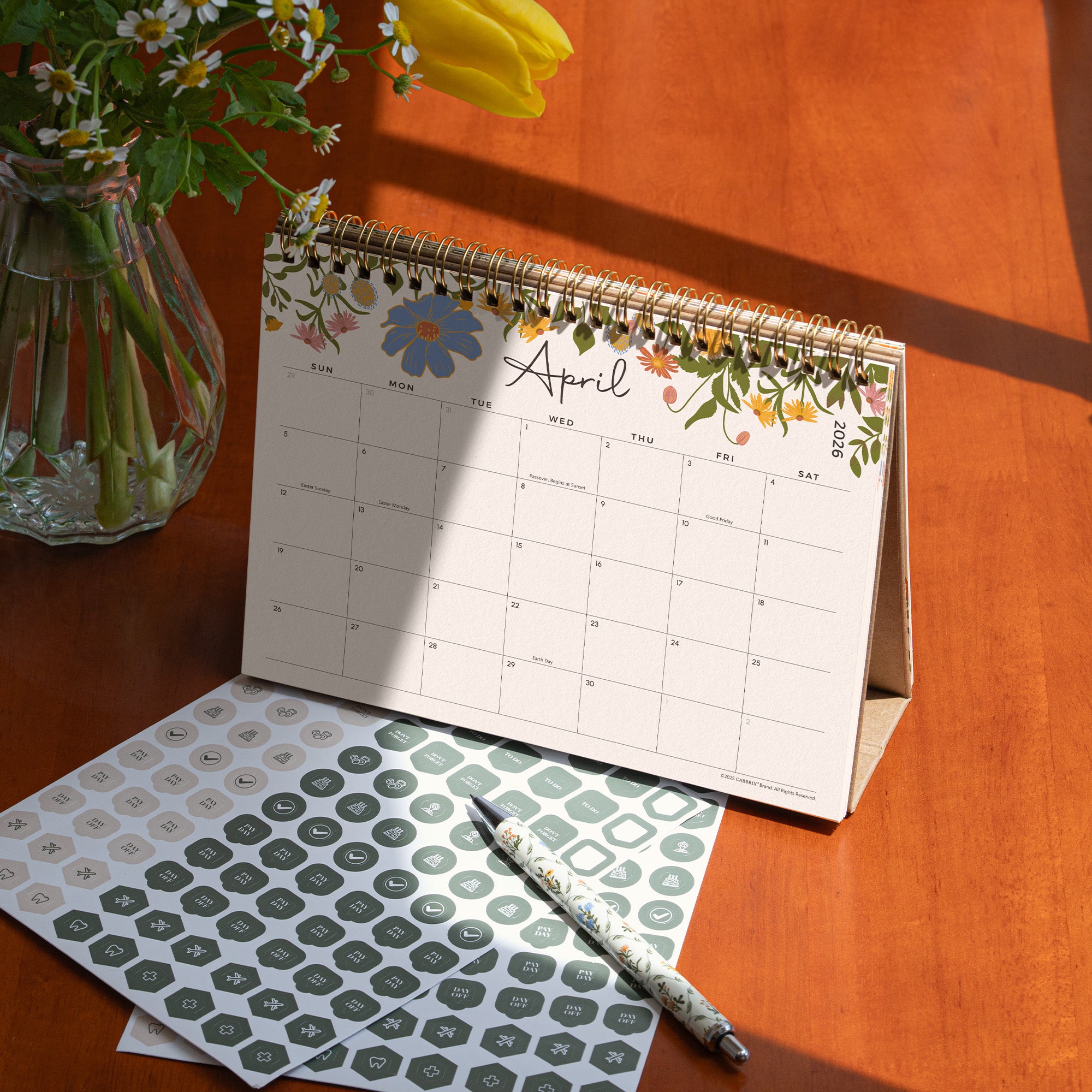 Floral-themed calendar open to April on a wooden surface with stationery items.