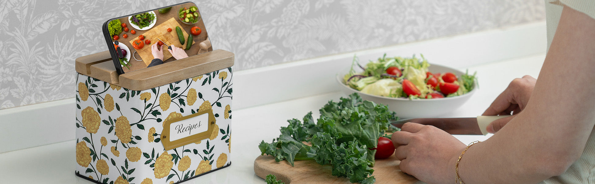 Person preparing a salad with a phone displaying a recipe above, on a kitchen counter.