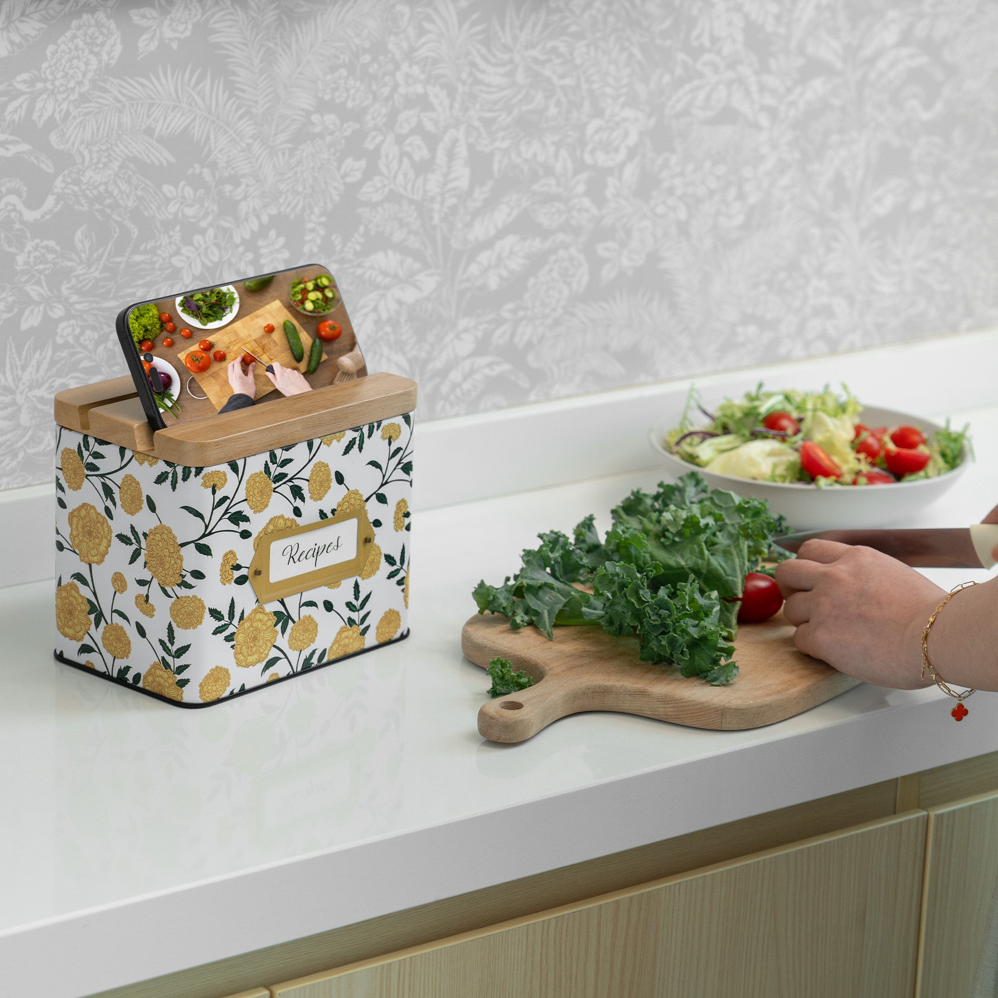 Kitchen counter with floral-patterned recipe box, cutting board with greens, and salad bowl.