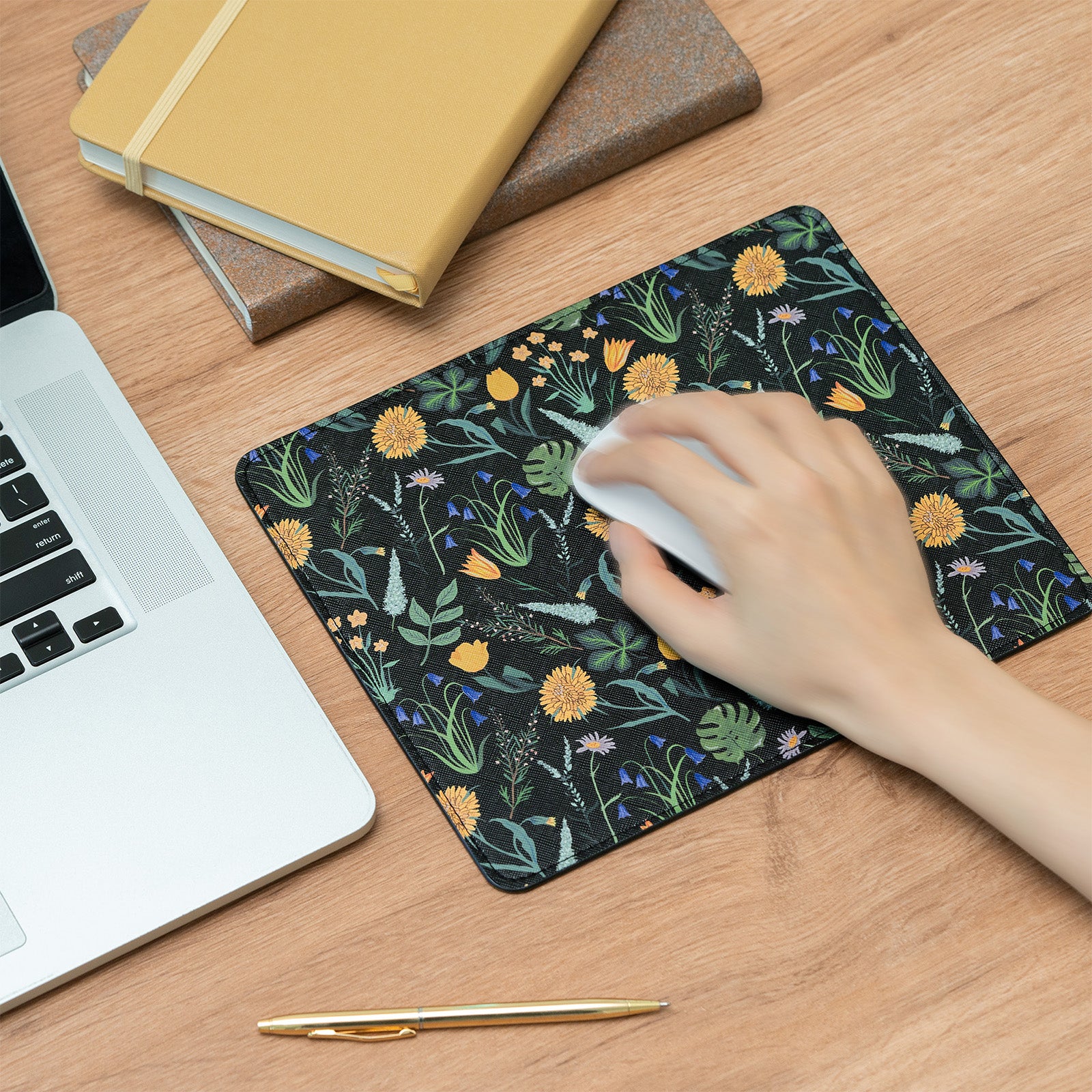 Hand using a mouse on a floral mouse pad with a laptop and notebooks in the background.