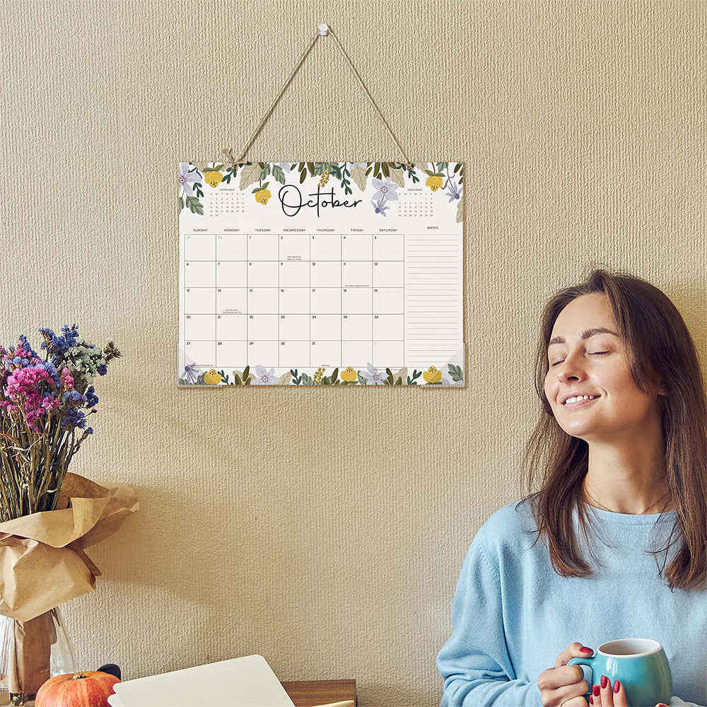 Woman sitting next to a floral-themed calendar on a wall, holding a mug.