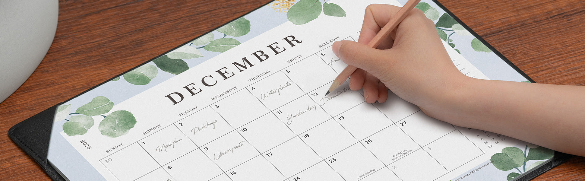 Hand holding a pen over a calendar with a decorative green leaf pattern on a wooden surface.
