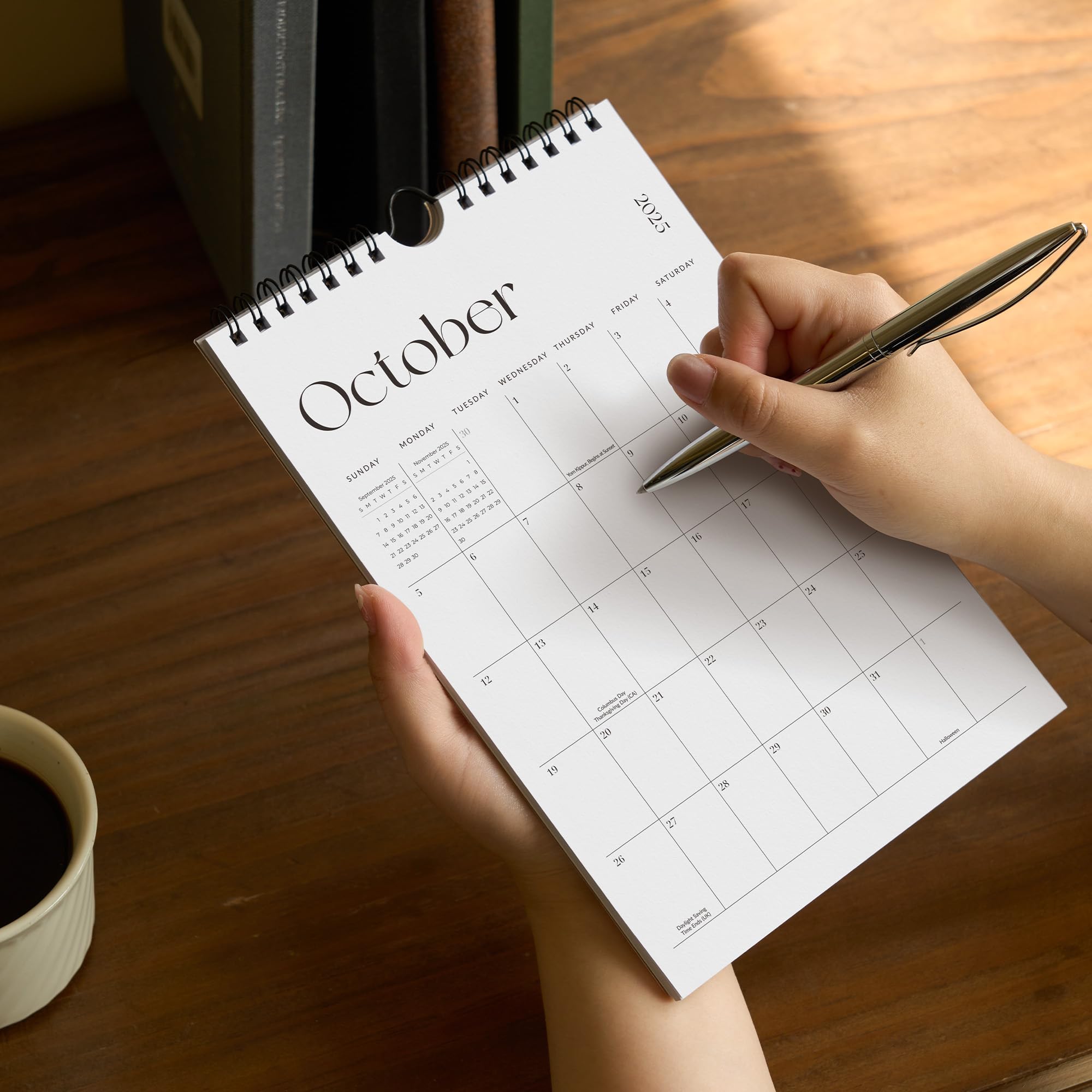 Person holding a spiral-bound calendar open to October on a wooden table with a pen.