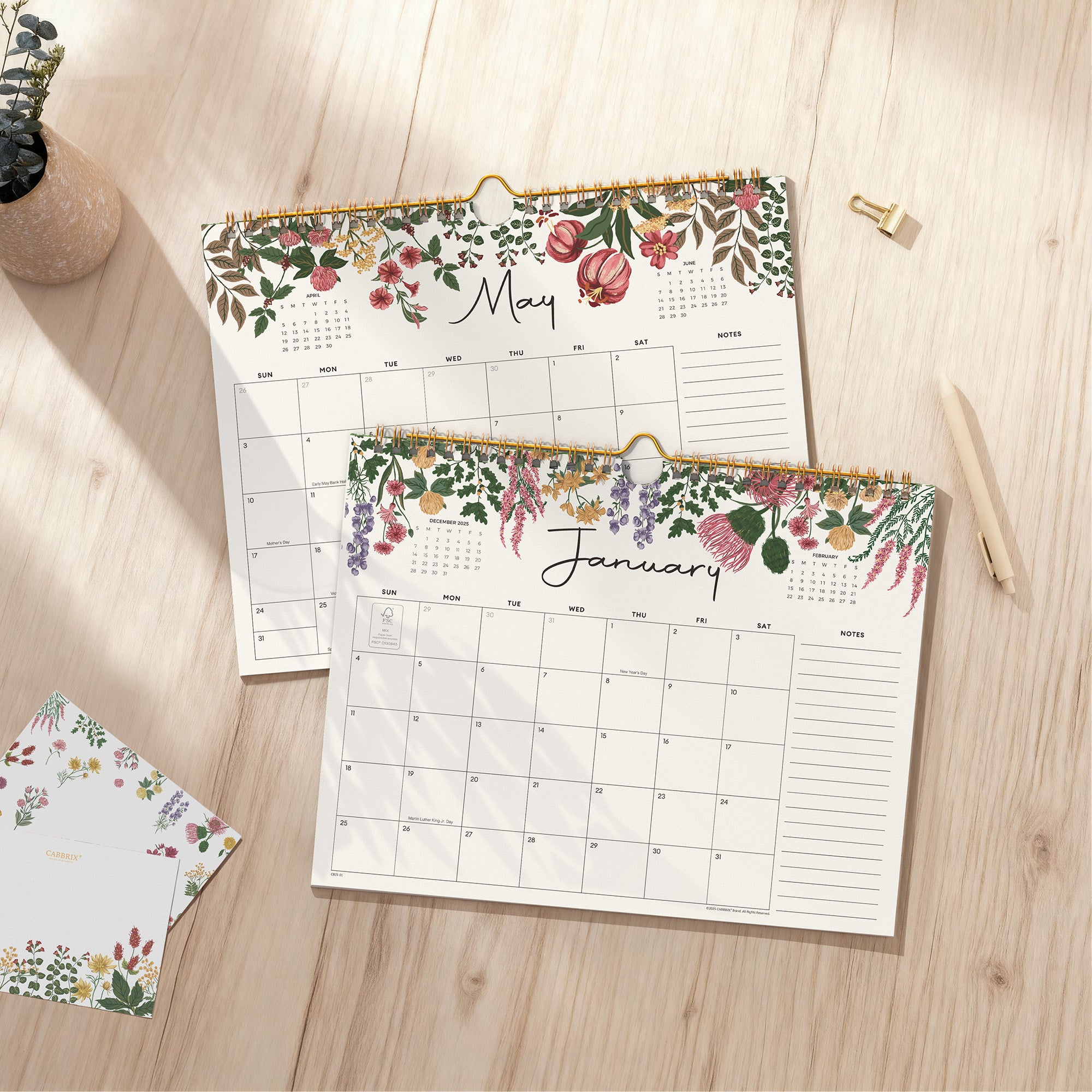Floral-themed calendar sheets on a wooden surface with a pen and small plant.