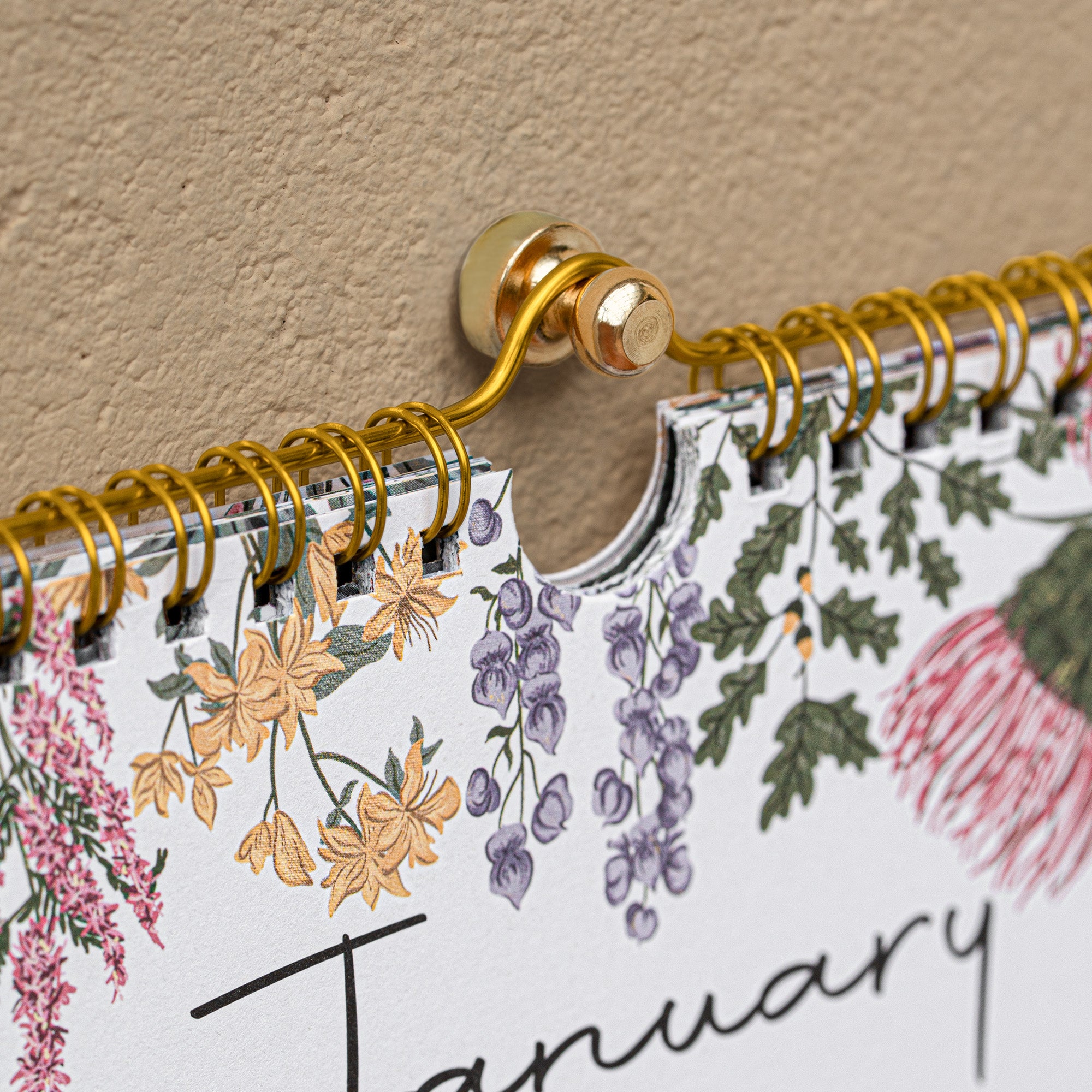 Floral-themed calendar with gold spiral binding against a beige wall.