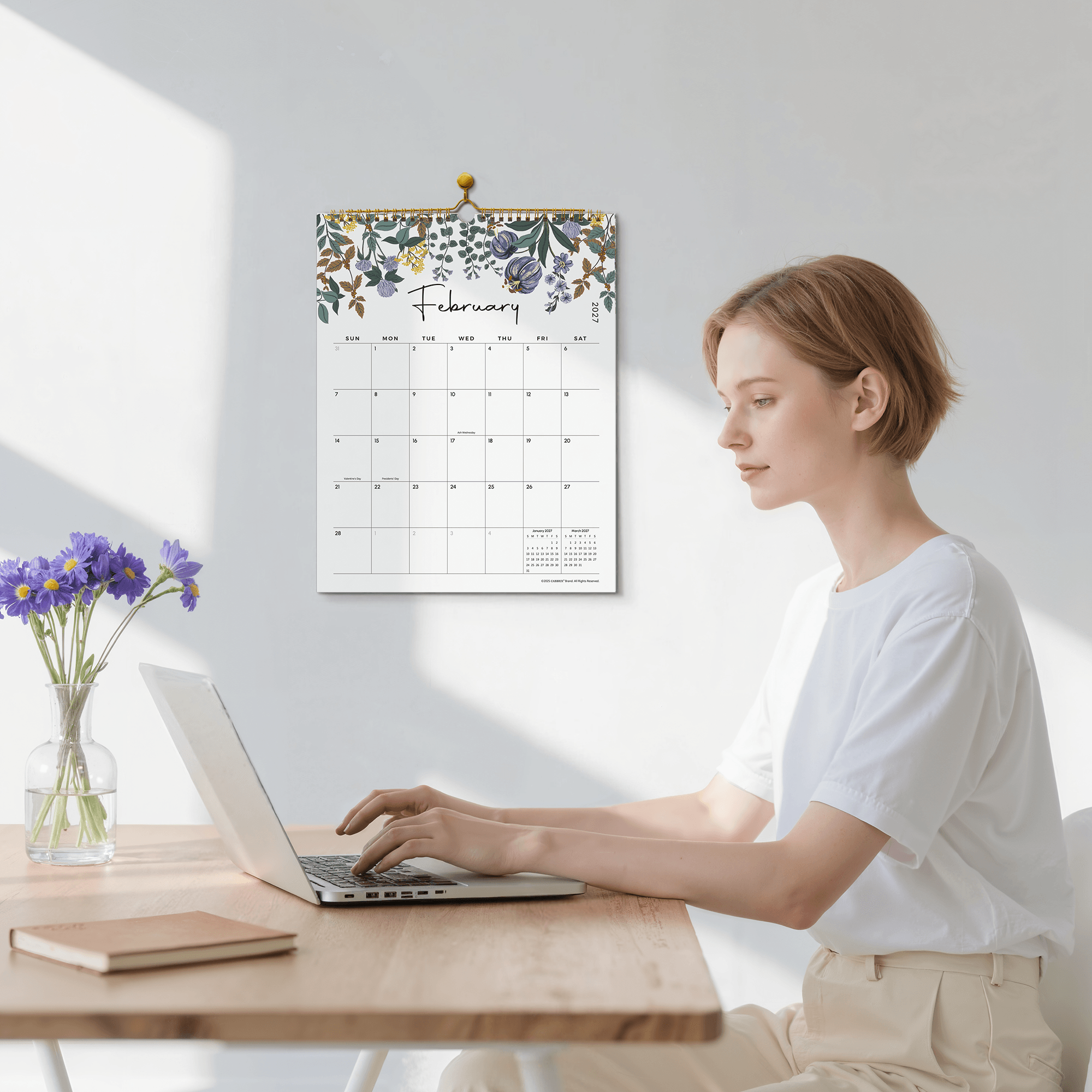 Woman sitting at a desk with a laptop, looking at a floral-patterned wall calendar.