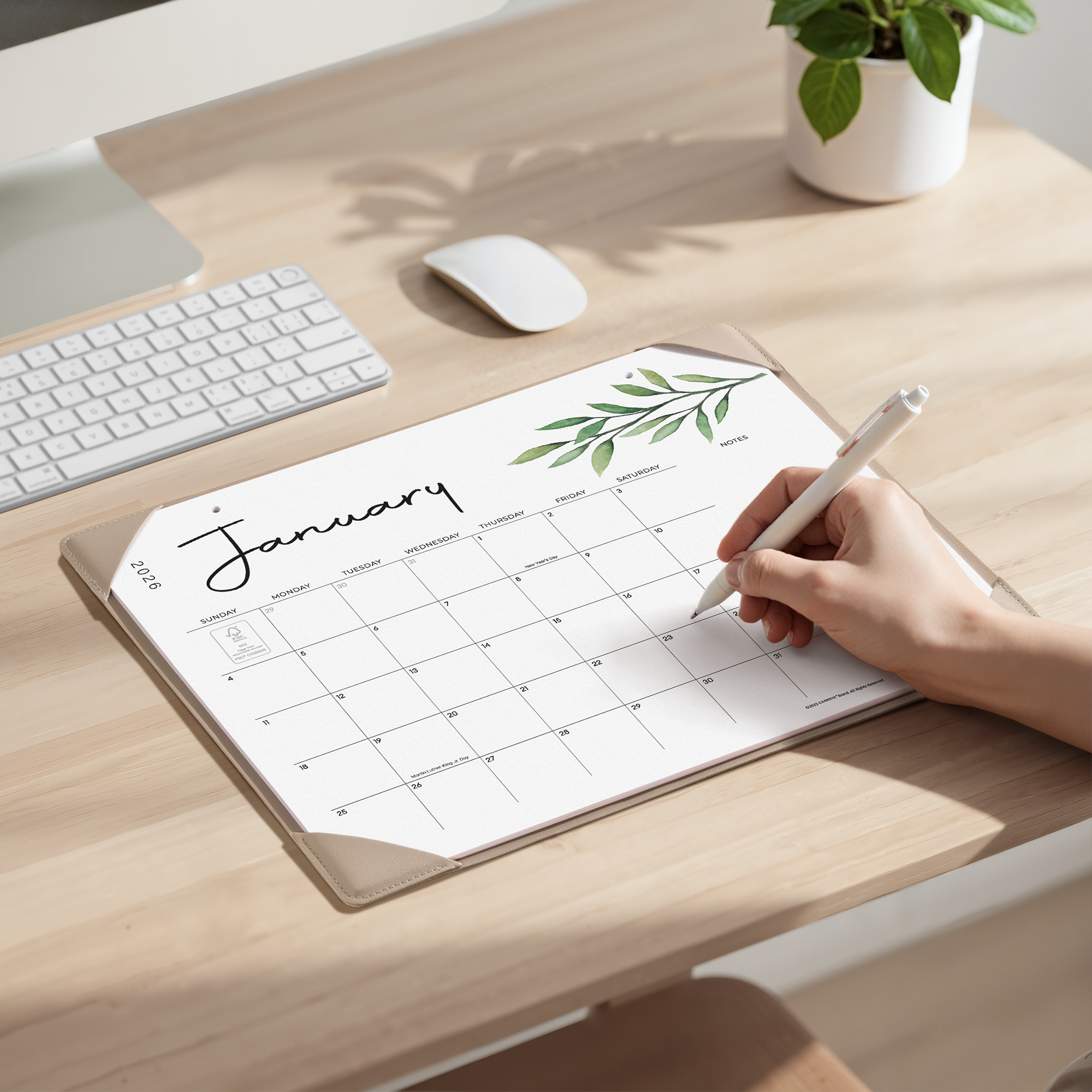 Person writing on a January calendar pad on a desk with a computer setup.