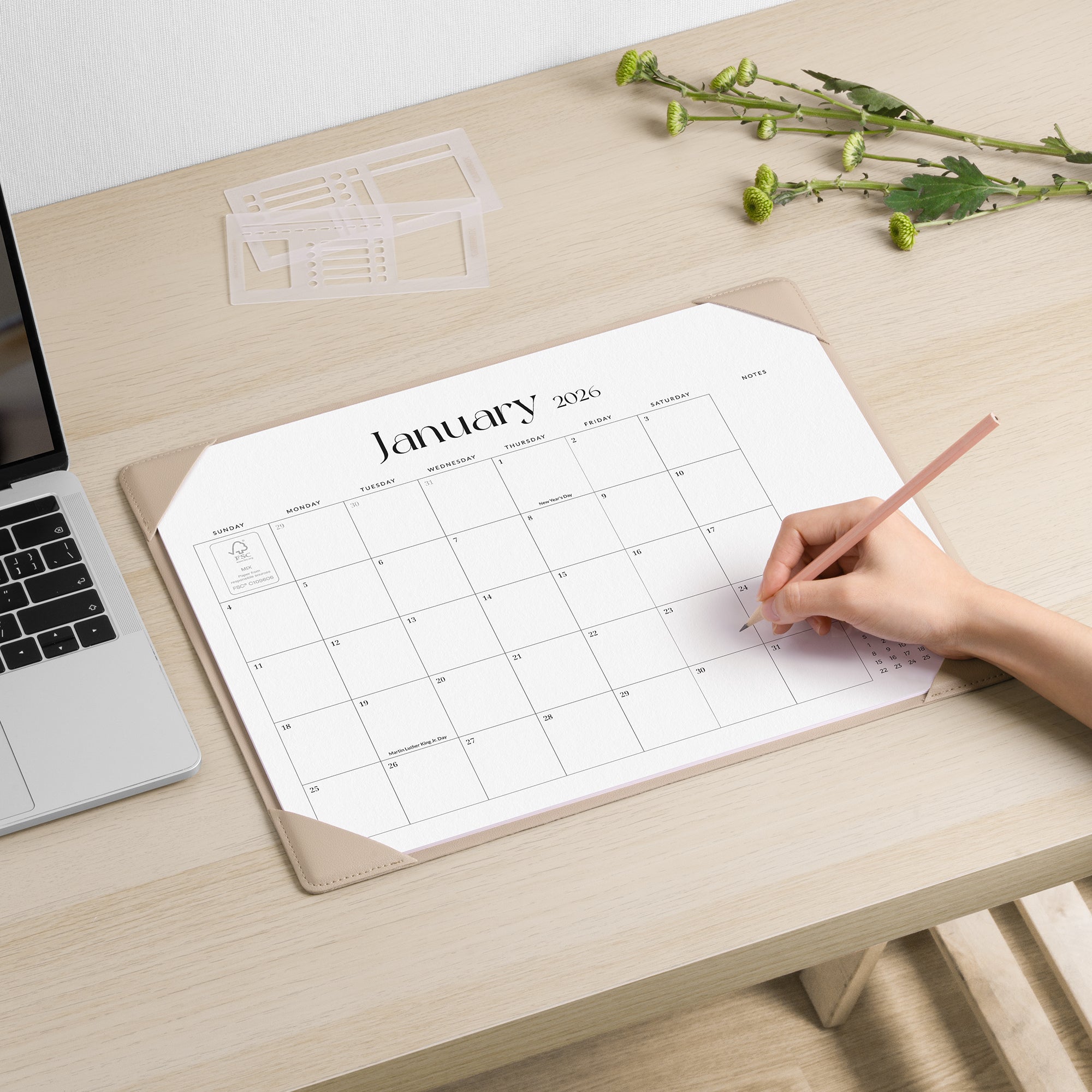Person using a pencil to mark on a January 2020 calendar on a desk with a laptop and flowers.