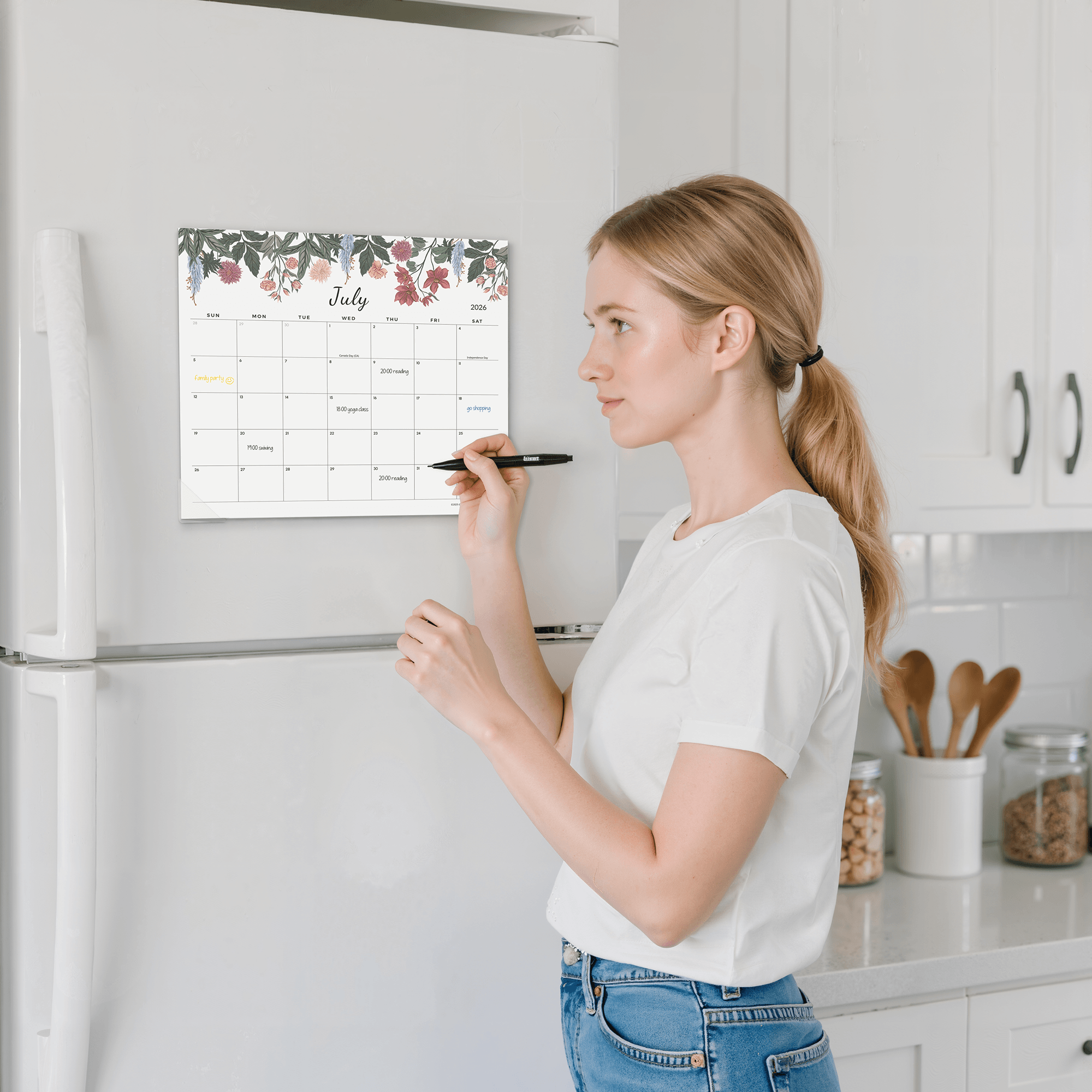 Woman writing on a floral calendar attached to a refrigerator in a kitchen.