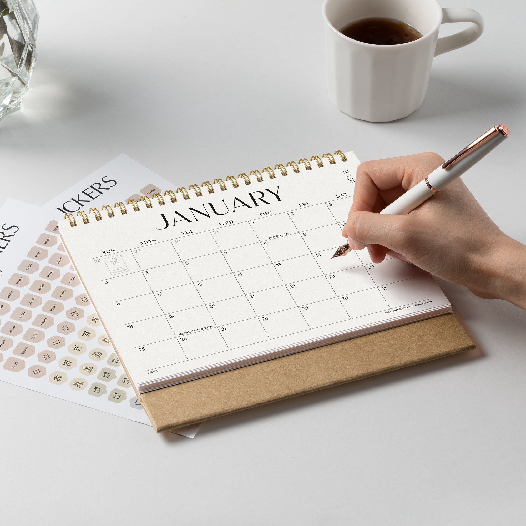 Hand holding a pen over a January calendar on a desk with a cup of coffee and a glass of water.