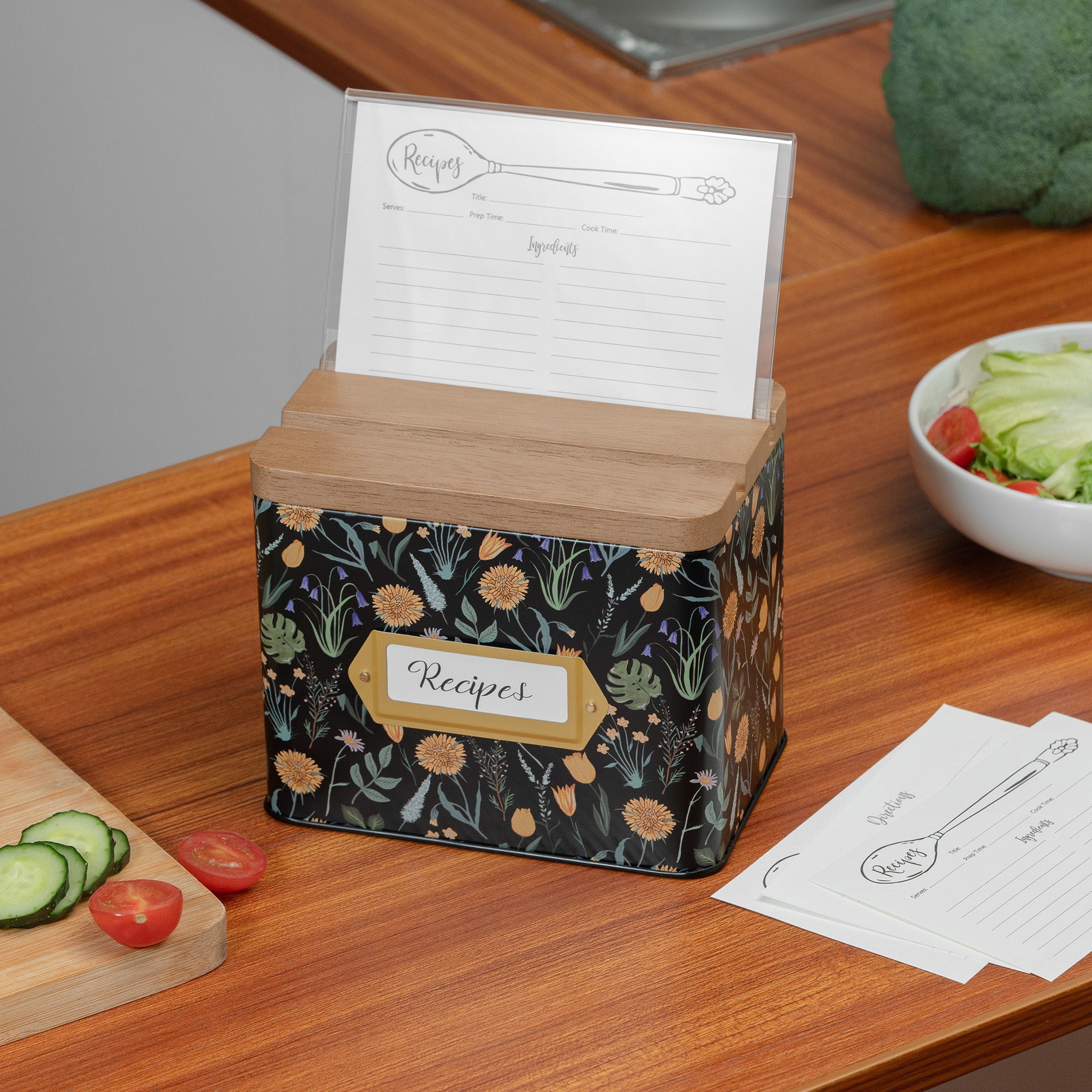 Floral-patterned recipe box on a kitchen counter with a cutting board and vegetables.