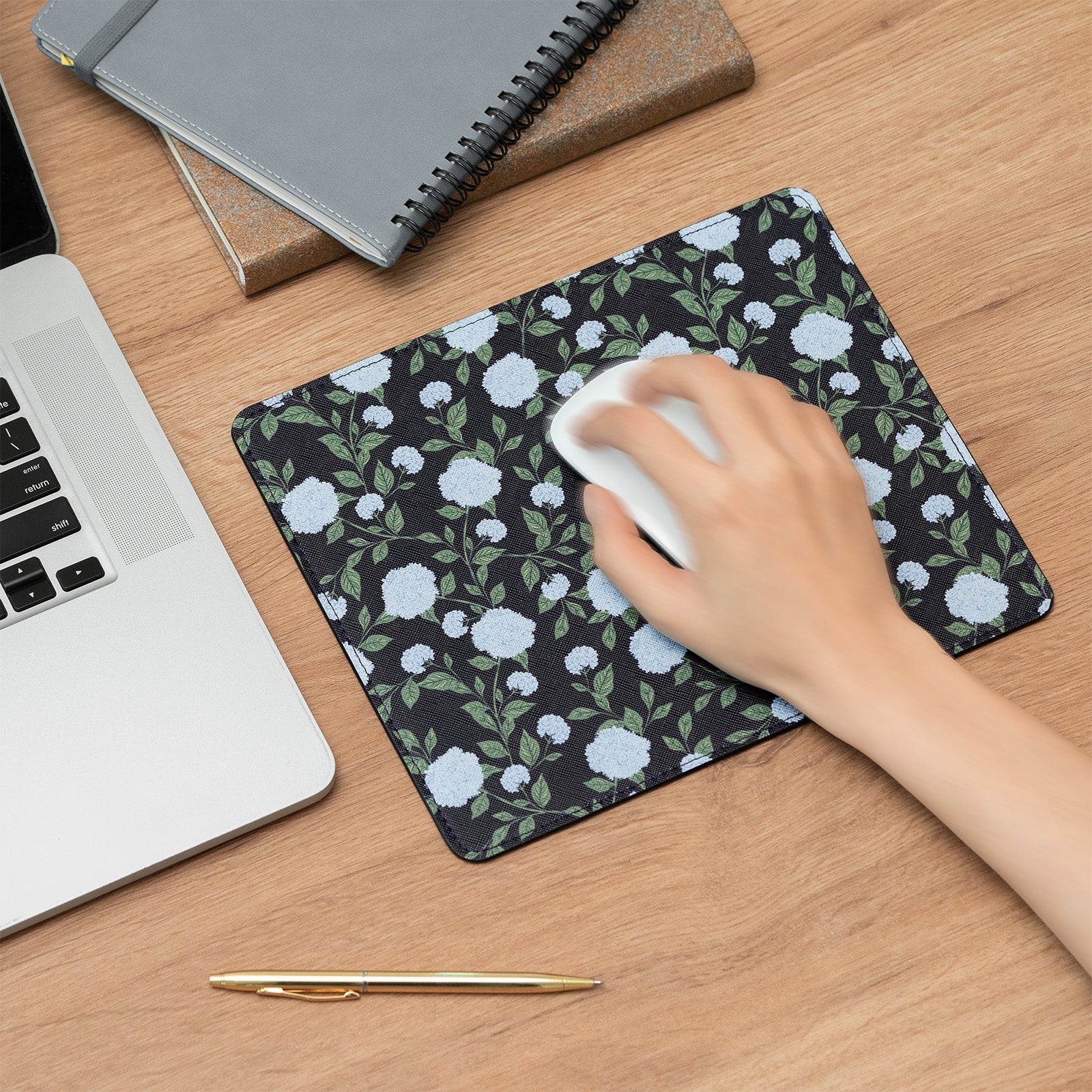 Hand using a white mouse on a floral mouse pad with a laptop and notebook in the background.