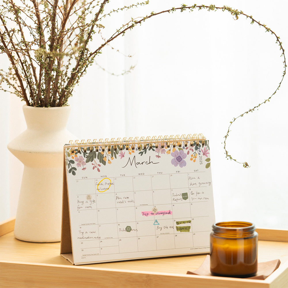 Calendar with floral design on a wooden surface next to a vase with dried plants and a candle.