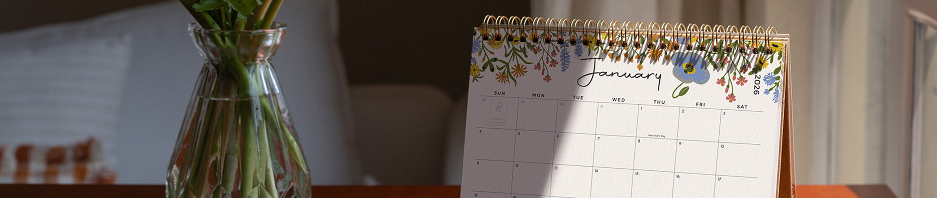 Calendar with floral design on a wooden surface, next to a vase with plants.